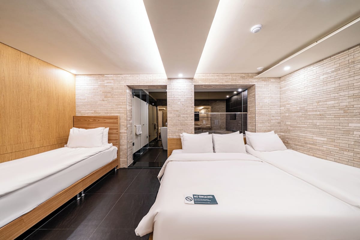 A modern hotel room with a large bed, a smaller bed, a wooden headboard, a brick wall, and a no smoking sign.