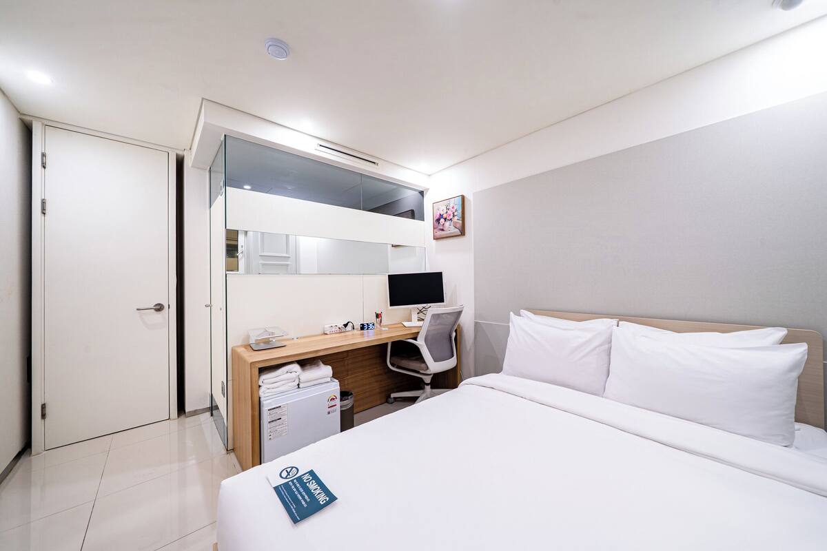 A hotel room with a bed, a desk with a computer, a chair, a wardrobe, and a small trash bin.