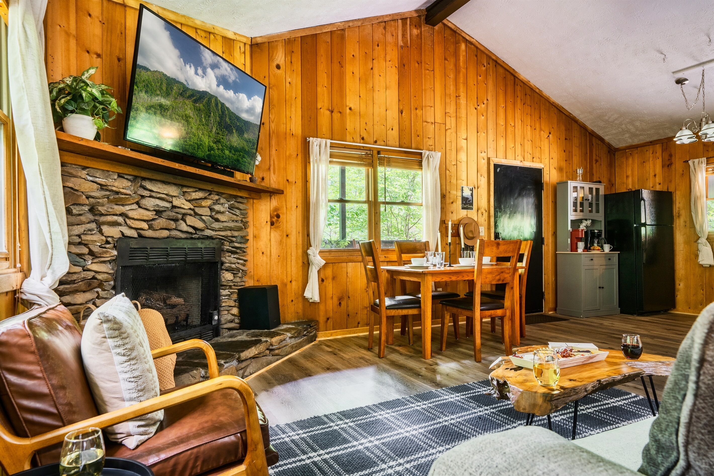 A cozy cabin with a stone fireplace, wooden walls, a dining area, and a kitchen with a refrigerator.