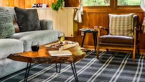 Um quarto acolhedor com piso de madeira, um sofá, uma poltrona, uma mesa de centro com uma bandeja de petiscos e uma taça de vinho, e uma janela com vista para o verde.