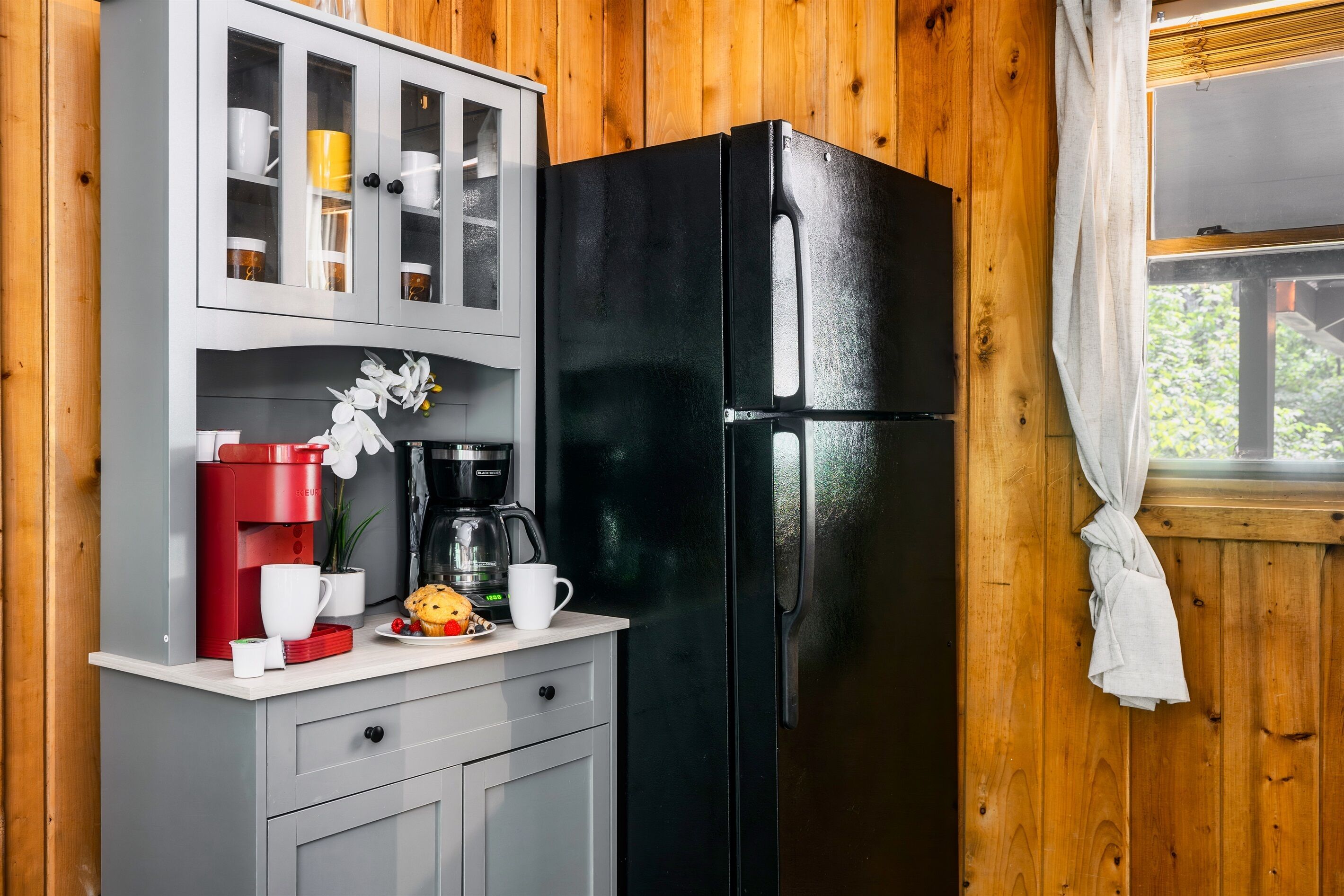 A kitchen with a black refrigerator, a coffee maker, and a cabinet with cups and a plant.