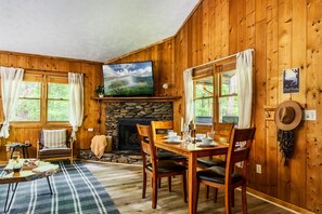 L’intérieur chaleureux d’un chalet avec une cheminée en pierre, des murs en bois, une table à manger dressée pour quatre personnes et un téléviseur fixé au-dessus de la cheminée.