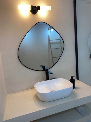 Un baño con un lavabo blanco, grifo negro y un espejo grande con marco negro. Hay una lámpara sobre el espejo y un dispensador de jabón negro en el lavabo.