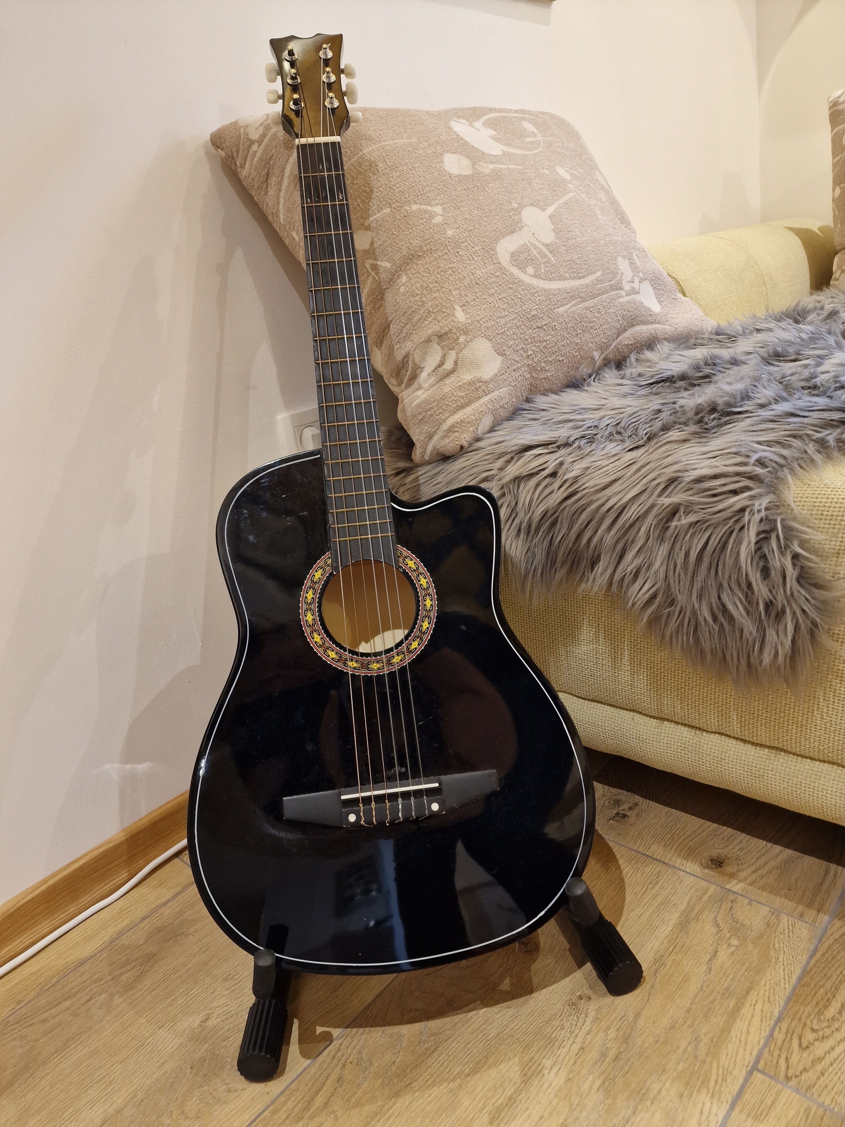 A black acoustic guitar with a decorative rosette, placed on a wooden floor next to a sofa with a patterned cushion.