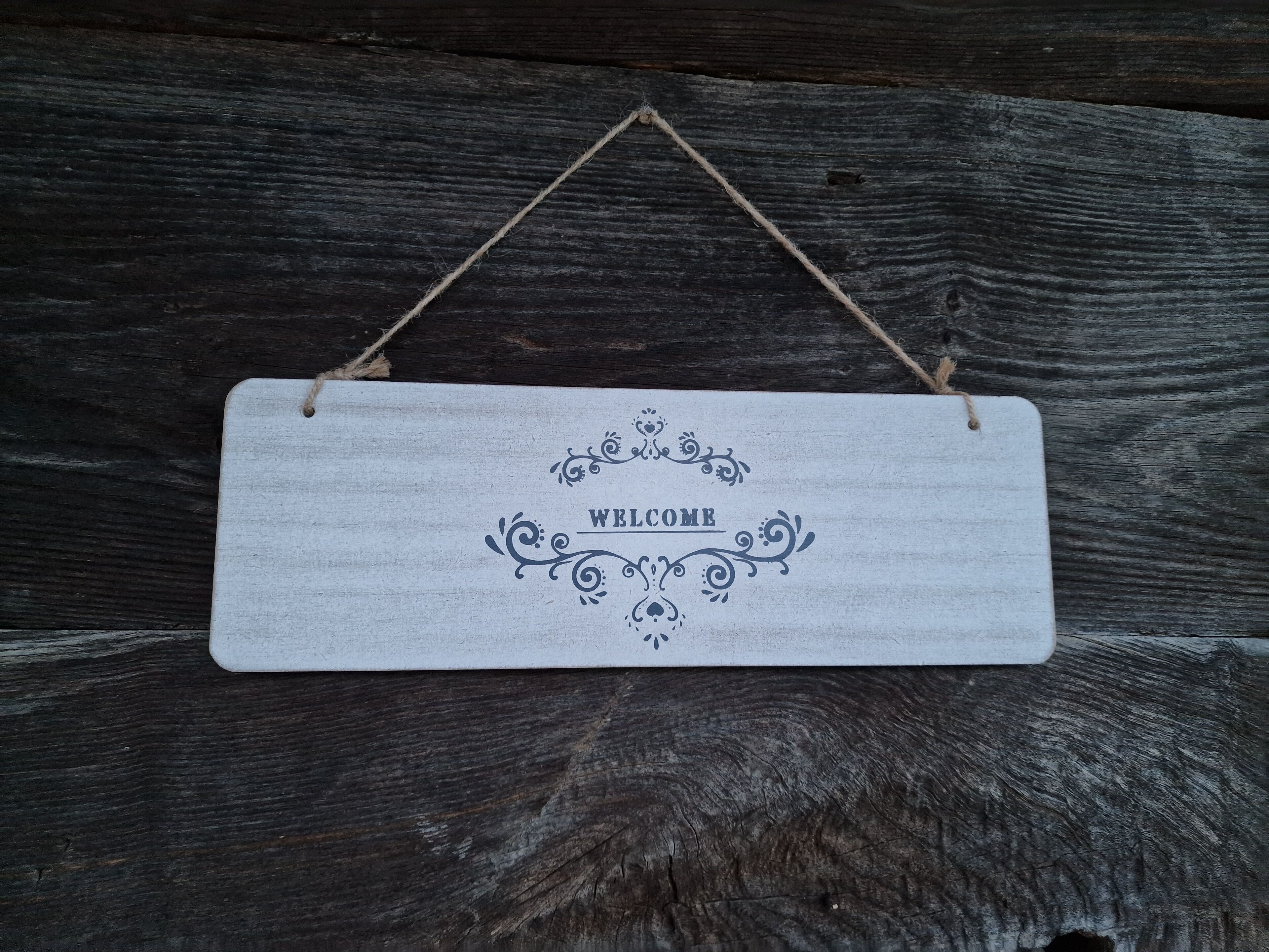Une enseigne rectangulaire en bois portant le message « BIENVENUE », suspendue sur un fond en bois foncé.
