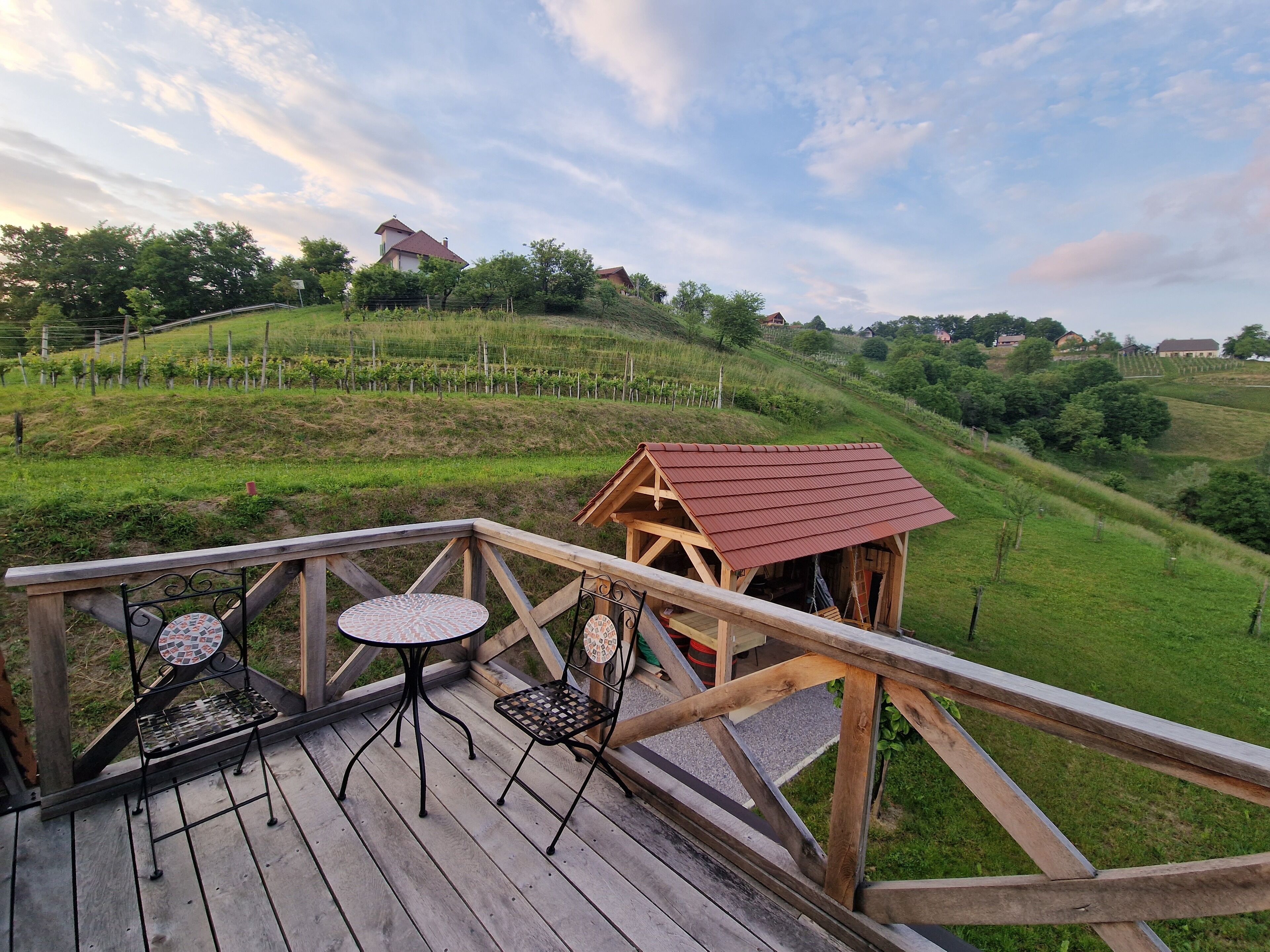 A wooden deck with a table and chairs, overlooking a vineyard and a hill with houses.