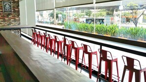 A seating area with red chairs and a long bench, large windows with a view of the street and greenery, and a brick wall with a framed picture.
