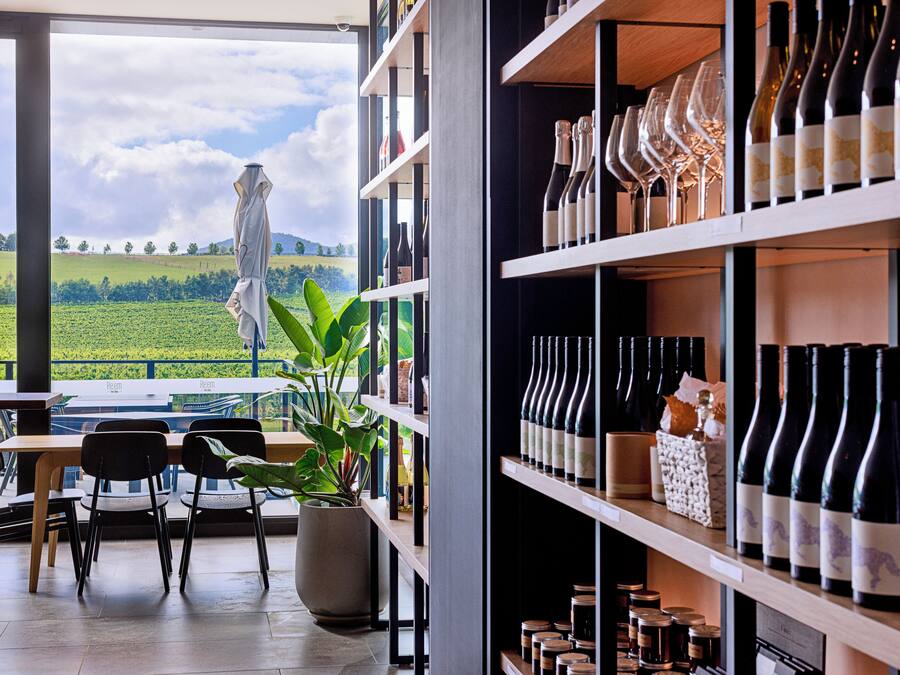 A restaurant with a view of a green field, a table with chairs, and shelves with wine bottles and glasses.