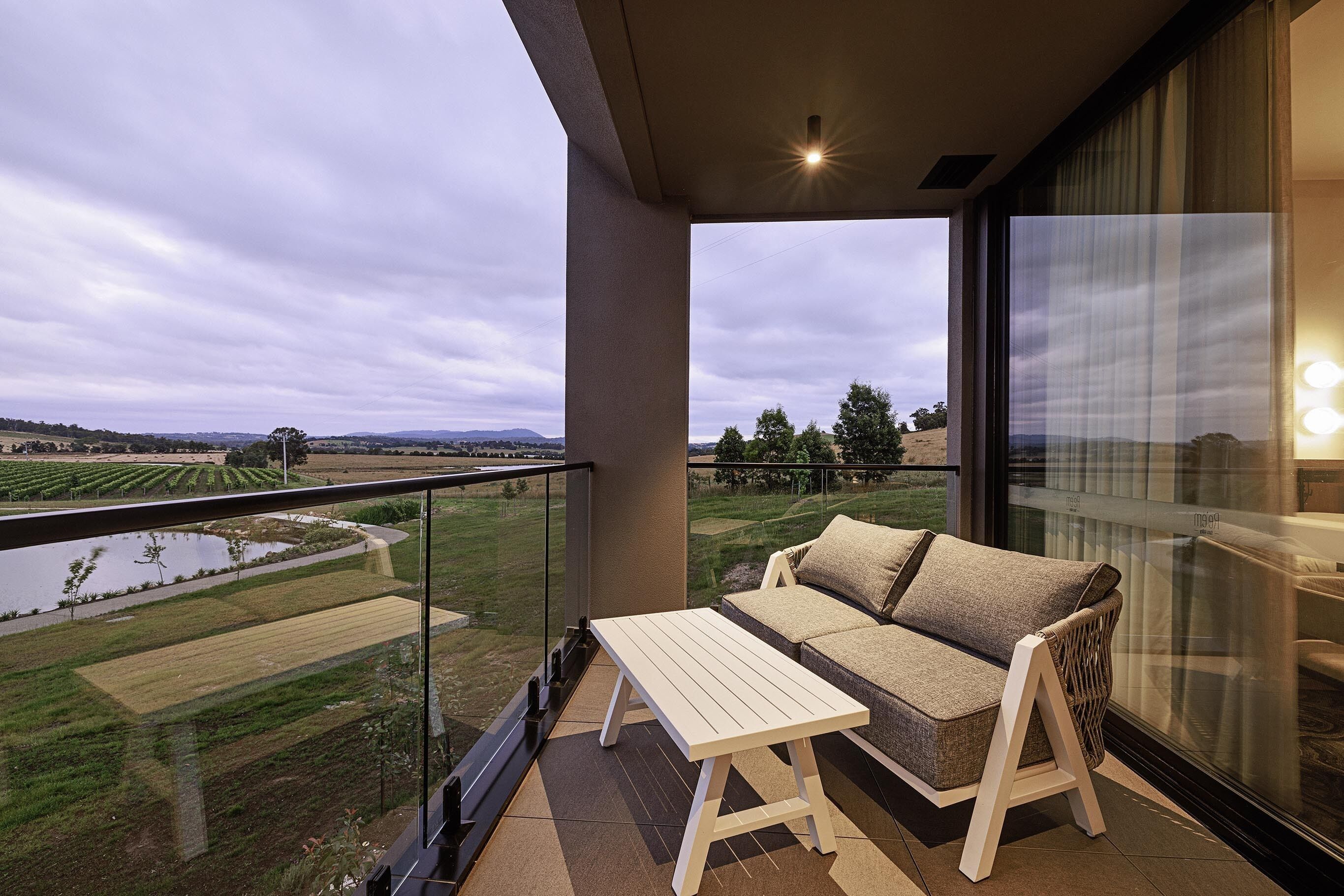 A balcony with a sofa and table, overlooking a vineyard and a body of water.