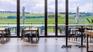 Un restaurante con grandes ventanales que ofrecen vista a un campo verde y colinas. Mesas y sillas están dispuestas en el interior.