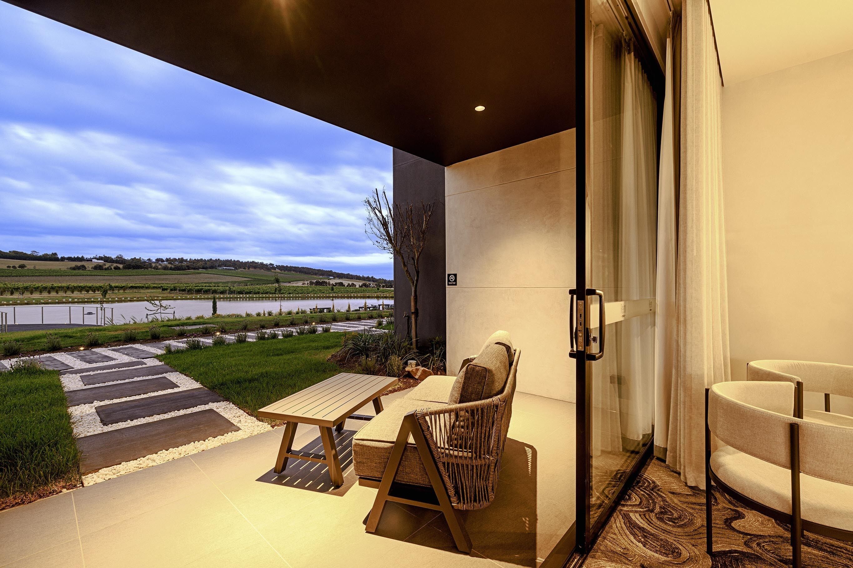 Eine moderne Außenpatio mit Tisch und Stühlen, eine Glastür, die zu einem Raum führt, und Blick auf einen See und Felder.