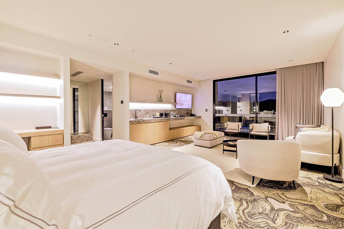 Habitación de hotel con una cama grande, un escritorio, una silla, un sofá, una televisión y vistas de la ciudad de noche.