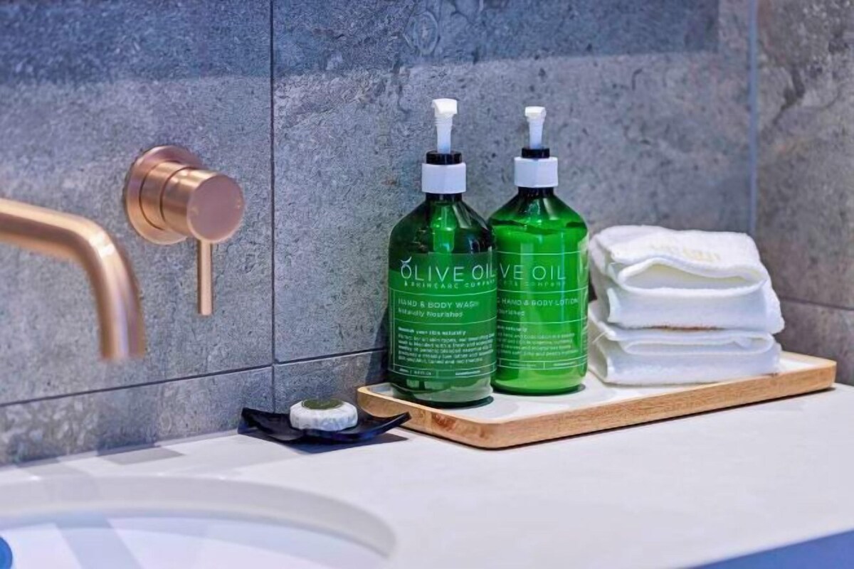 Un baño con un grifo moderno, dos botellas de aceite de oliva y una pila de toallas blancas sobre una bandeja de madera.