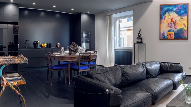 A modern kitchen with dark cabinetry, a dining area with blue chairs, and a black leather sofa. - Luxury modern apartment (Roma)