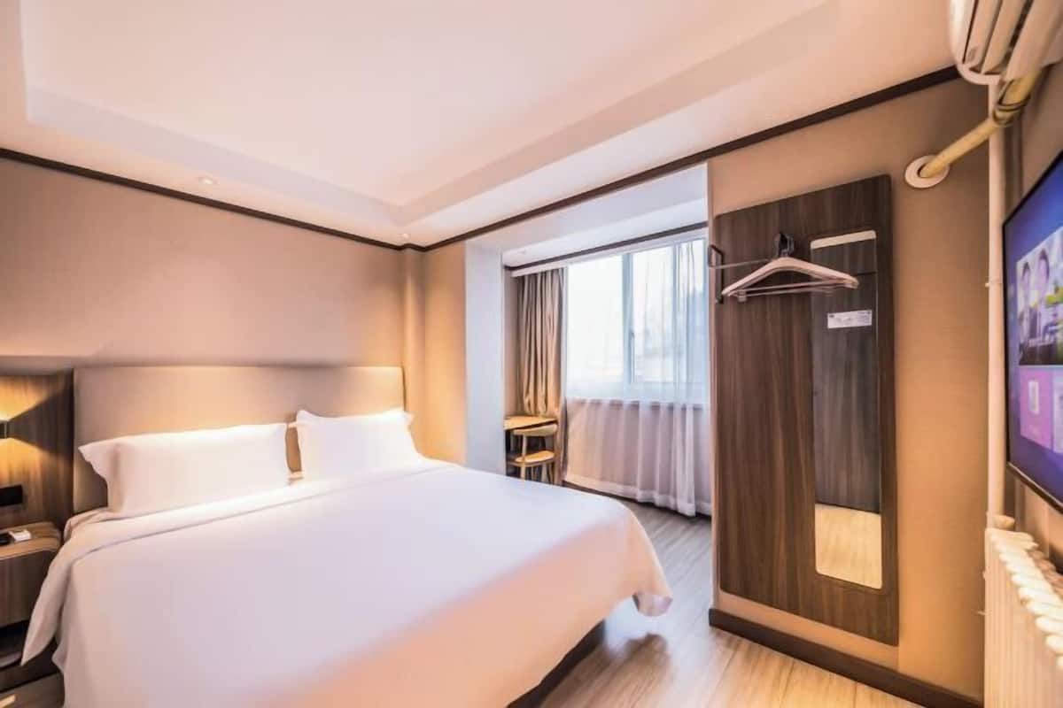A hotel room with a large bed, bedside tables, a TV, a mirror, and a window with curtains.