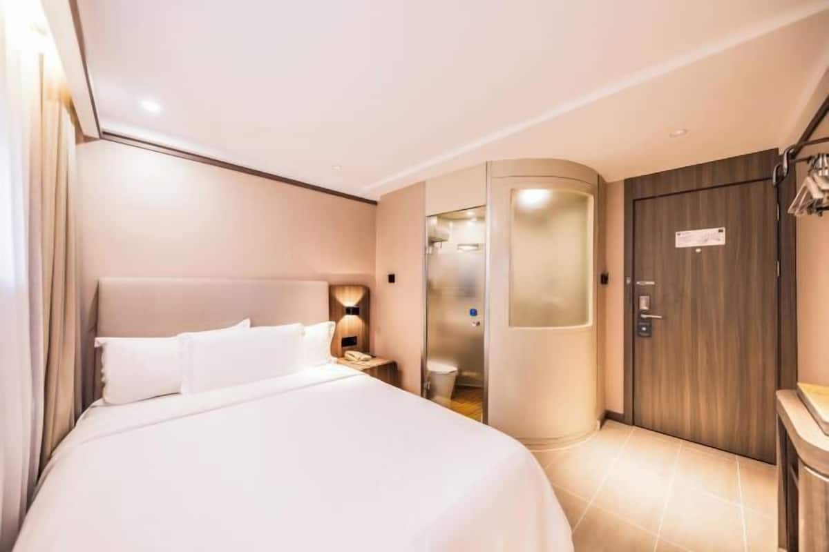 A modern hotel room with a large bed, a bedside table with a lamp, a wardrobe, and a shower area.