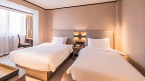 Habitación de hotel con dos camas, una mesita de noche con lámpara, una silla y un amplio ventanal con cortinas.