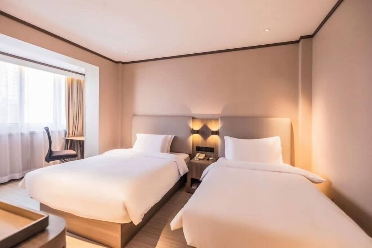 A hotel room with two beds, a bedside table with a lamp, a chair, and a large window with curtains.