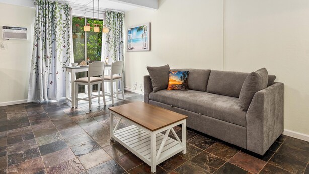 Ein Wohnzimmer mit grauem Sofa, einem Holztisch, einem Esstisch mit Stühlen und Blick ins Freie durch ein Fenster mit Vorhängen.