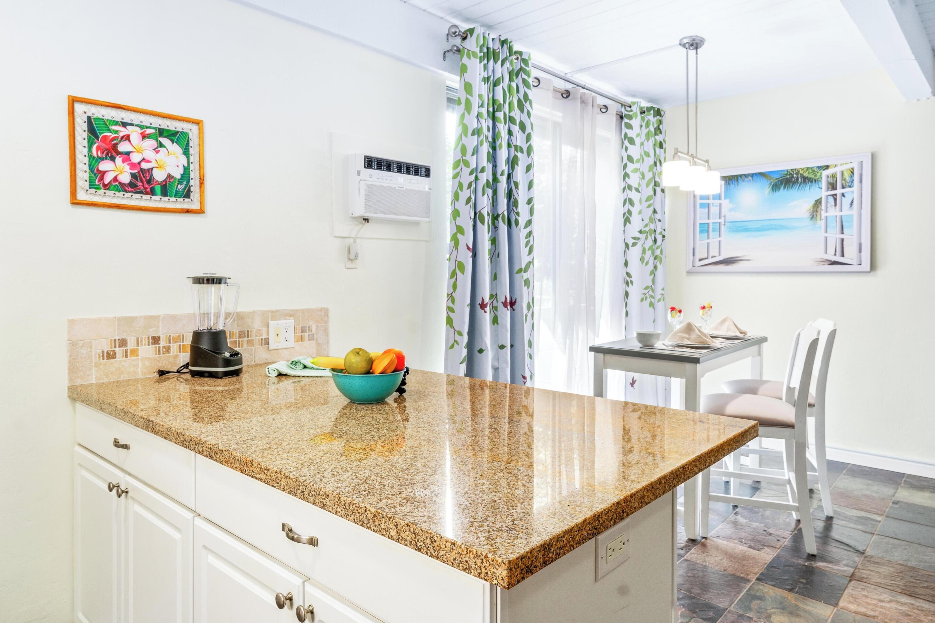 A kitchen with a granite countertop, a fruit bowl, a blender, and a table with a chair. There is a painting on the wall and a window with curtains.