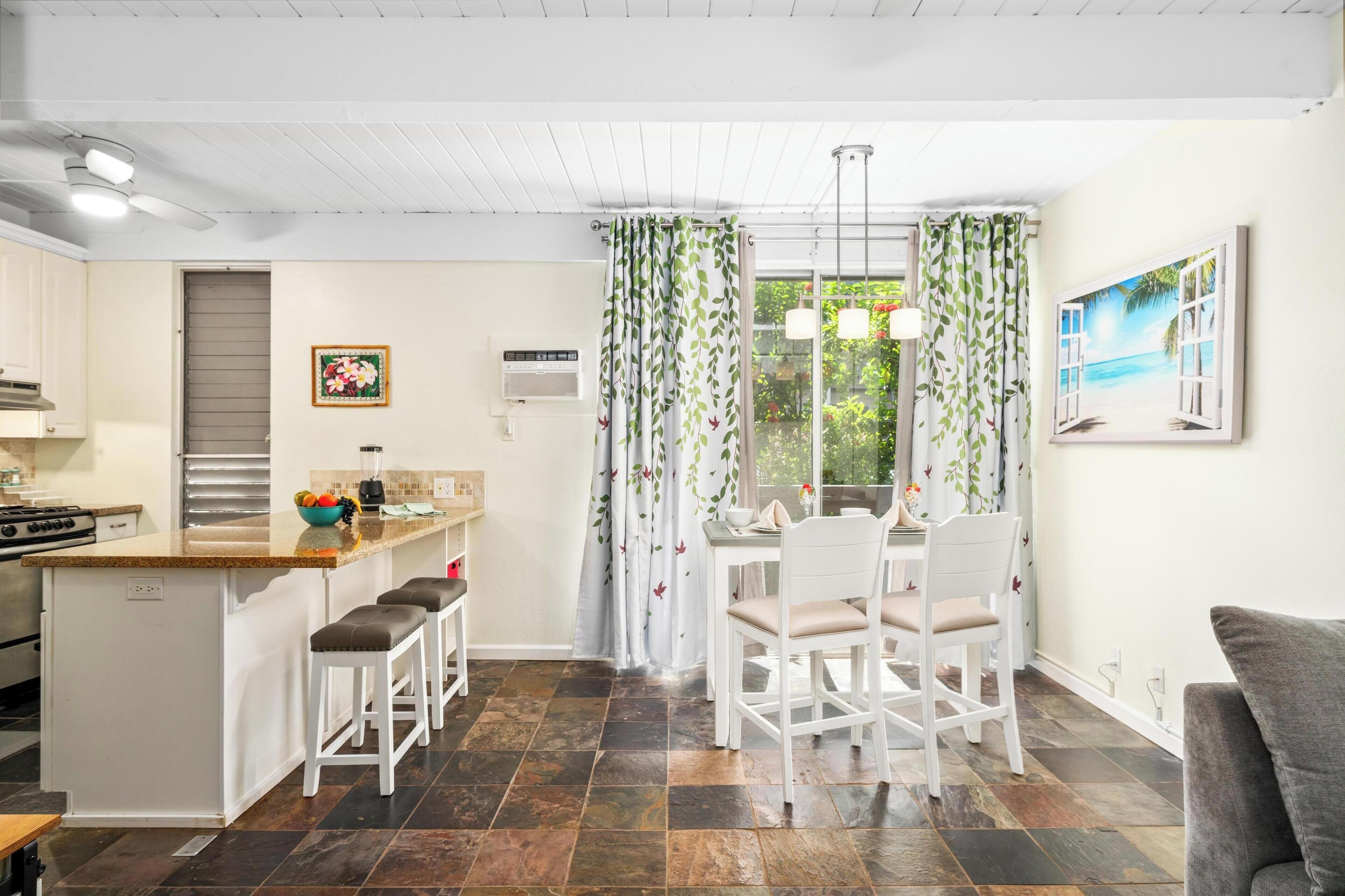 A modern kitchen with a central island, white bar stools, a dining area with a white table and chairs, and a large window with curtains.