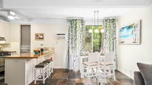 Eine moderne Küche mit einer zentralen Kochinsel, weißen Barhocker, einem Essbereich mit weißem Tisch und Stühlen sowie einem großen Fenster mit Vorhängen.