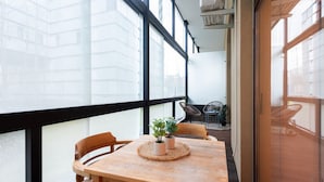 A balcony with a wooden table and chairs, a potted plant, and a view of the cityscape through large windows.