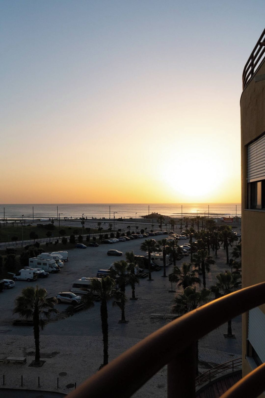 A parking area with vehicles and palm trees, overlooking a beach and ocean at sunset.