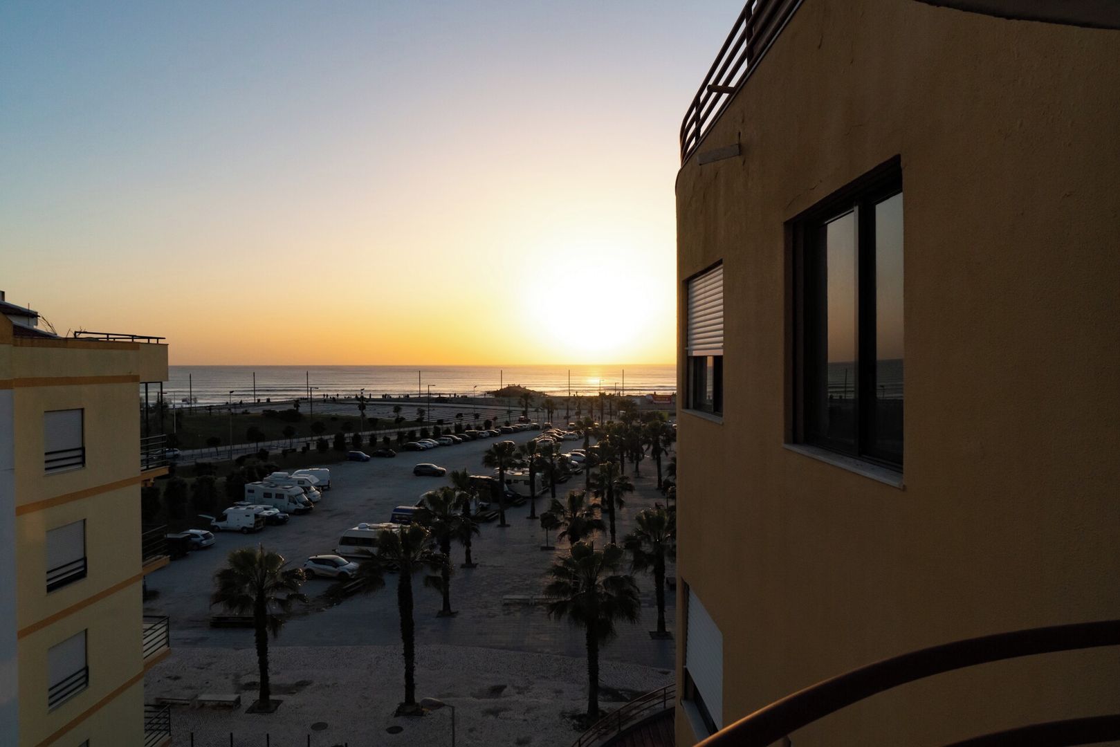 A view of a parking lot with several cars and palm trees, leading to a beach with a sunset in the background.