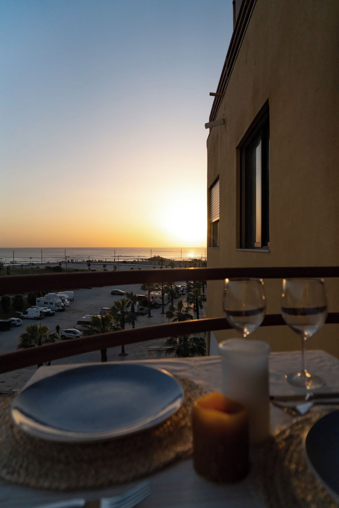 A balcony with a table set for two, overlooking a parking area and a beach.