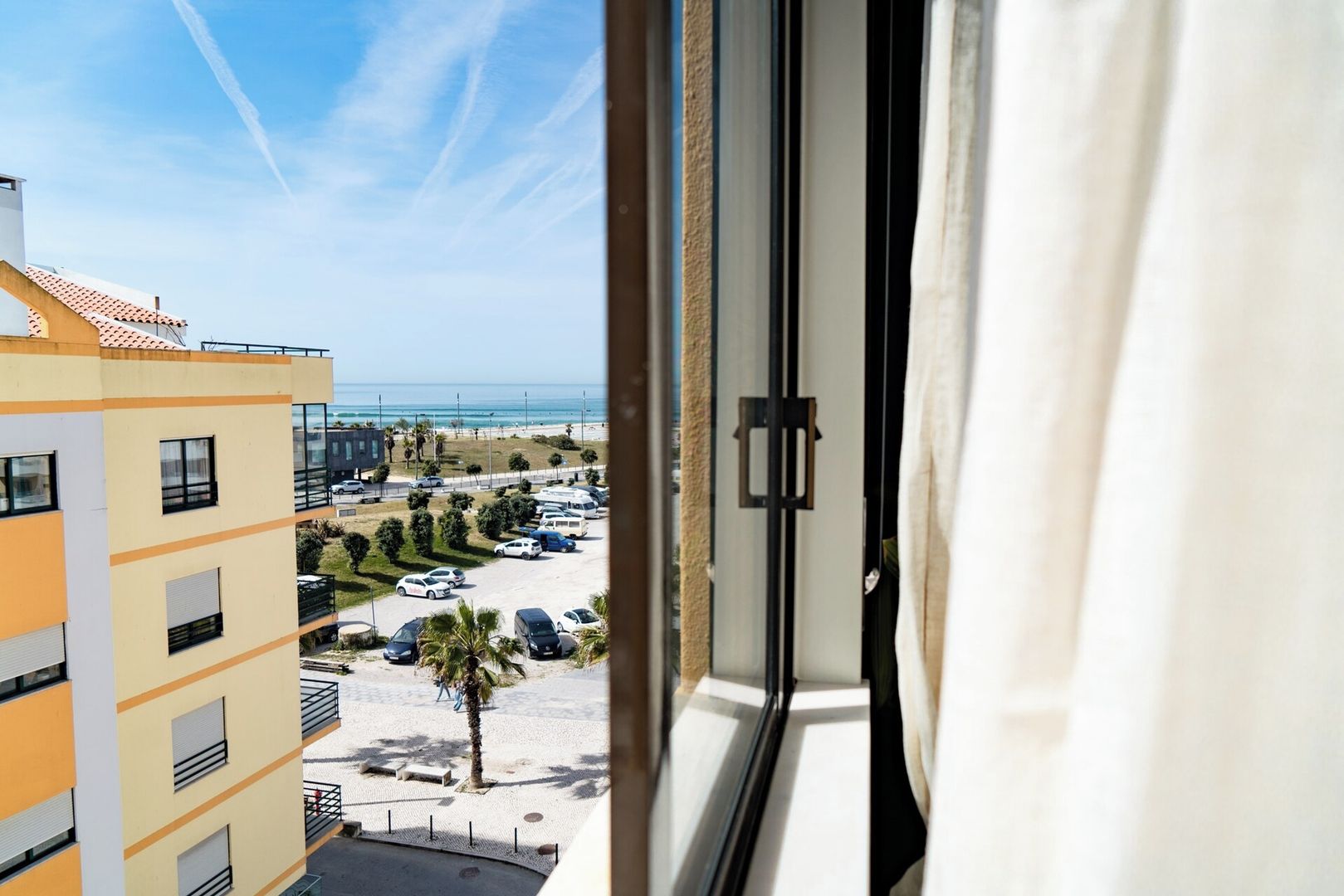 A view from a window overlooking a parking area with a building in the foreground and a beach in the background.