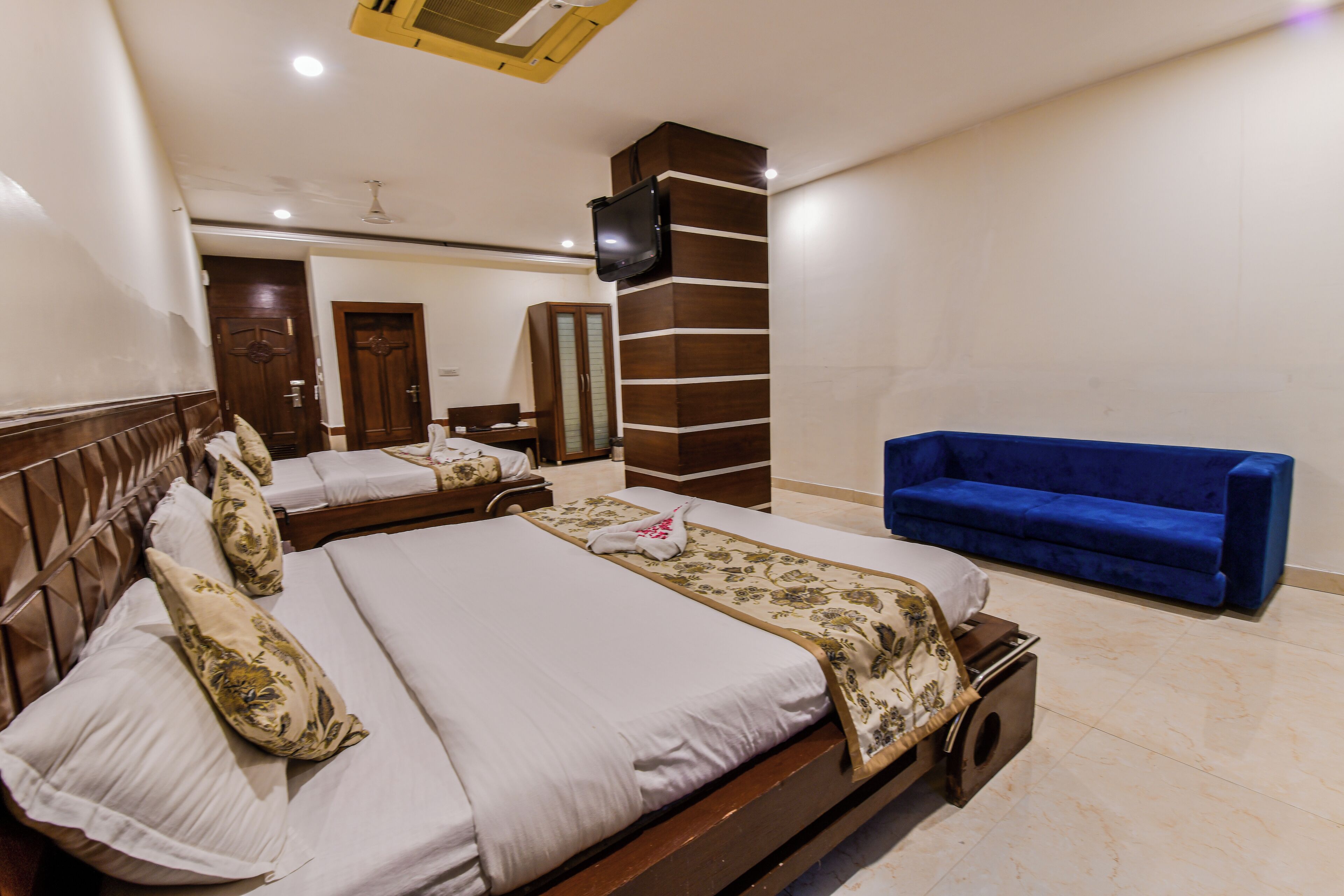 A hotel room with a large bed, a blue sofa, a TV mounted on a wooden pillar, and a wardrobe.
