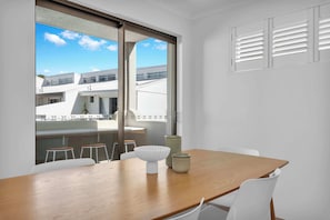 Uma sala de jantar moderna com mesa de madeira, cadeiras brancas e uma janela grande com vista para edifícios.