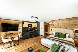 Une chambre d’hôtel moderne avec un plancher en bois, un coin repas comprenant une table et des chaises, une cuisine avec des meubles de couleur sombre et un grand lit agrémenté d’oreillers verts.