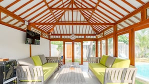A covered outdoor seating area with wooden benches and a table, a TV mounted on the wall, and a view of a swimming pool. - Villa Vie (Lembongan Island)