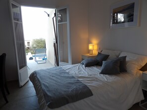 Une chambre à coucher avec un lit, des oreillers, une table de chevet, une lampe et une porte ouverte donnant sur un balcon.