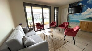 A modern living room with a grey sofa, red chairs, a small table, and a view of a balcony with outdoor seating.