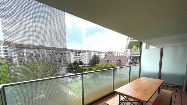 A balcony with a wooden table and benches, a mesh safety screen, and a view of residential buildings.