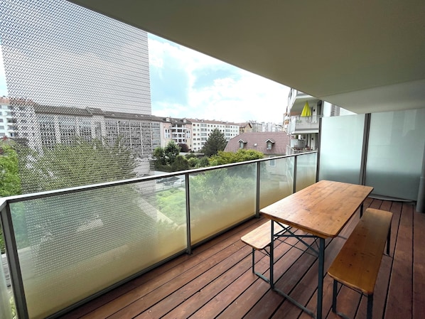 Ein Balkon mit Holztisch und Bänken, einem Drahtnetzgitter und Blick auf Wohnhäuser.