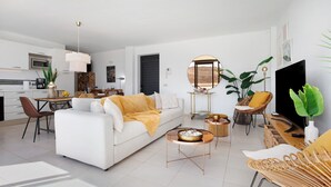 A modern living room with a sofa, coffee table, and TV. There is a dining area with chairs and a table.