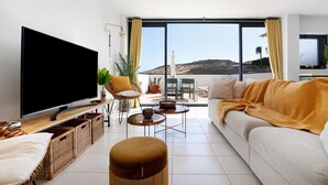 A modern living room with a large flat-screen TV, a sofa, a coffee table, wicker baskets, and a view of a hill through large windows.