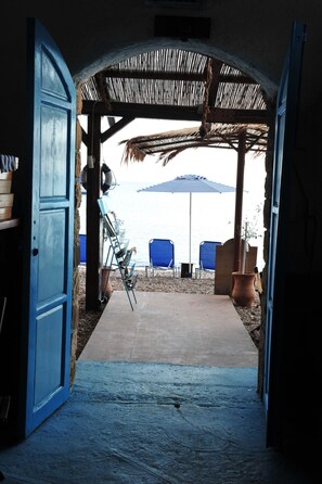 Une porte ouverte donne sur un espace extérieur abrité, doté d’un sol en béton, de chaises bleues, d’un parasol bleu et d’un toit de chaume rustique.