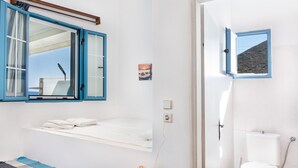 Une chambre avec un lit, une fenêtre avec des volets bleus, une petite table de chevet, une lampe fixée au mur et une vue sur la mer.