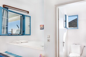 Une chambre avec un lit, une fenêtre avec des volets bleus, une petite table de chevet, une lampe fixée au mur et une vue sur la mer.