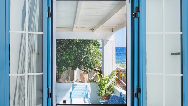 Une fenêtre ouverte, dotée d’un cadre bleu, offre une vue sur un balcon, un panier en osier, une chaise bleue et un pot de fleurs.