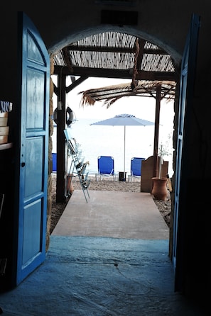 An open doorway leading to a covered outdoor area with a concrete floor, blue chairs, a blue umbrella, and a rustic thatched roof.
