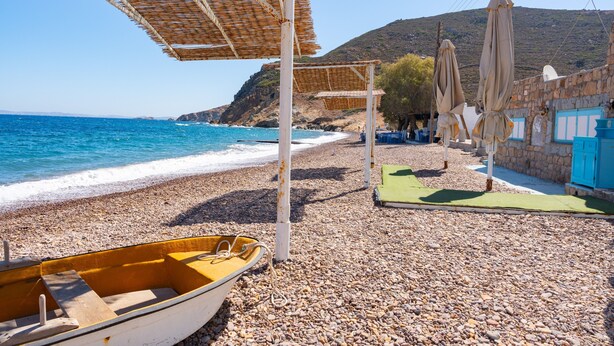 Een kiezelstrand met een boot, parasols en een stenen gebouw.