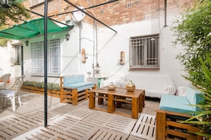 A courtyard with wooden furniture, a green umbrella, and a white wall with windows.