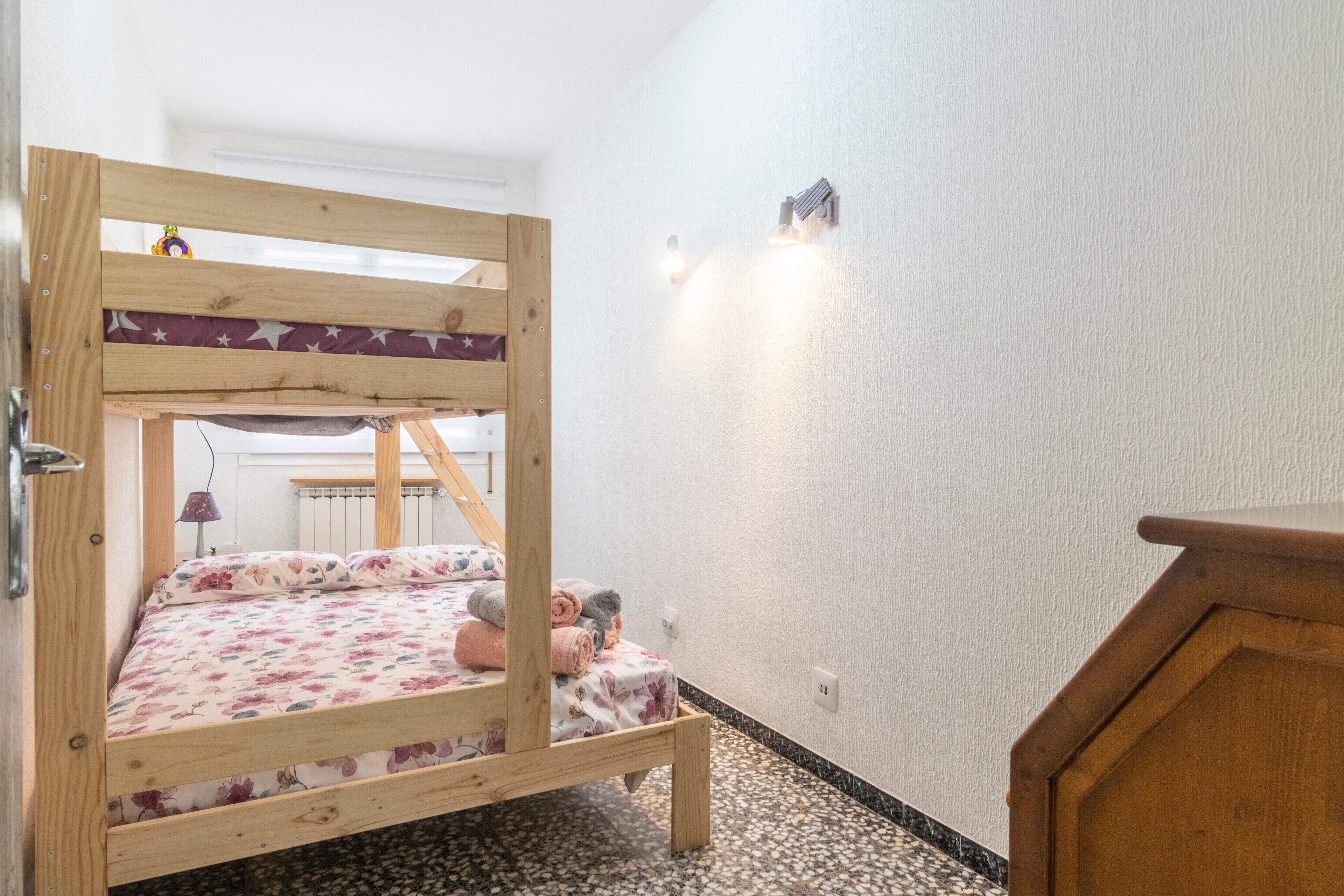 En køjeseng med blomstret dyne, et lille skrivebord med lampe, og en radiator i baggrunden.