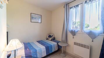 Een kleine kamer met een enkel bed, een raam, een radiator, een nachtkastje en een lamp.